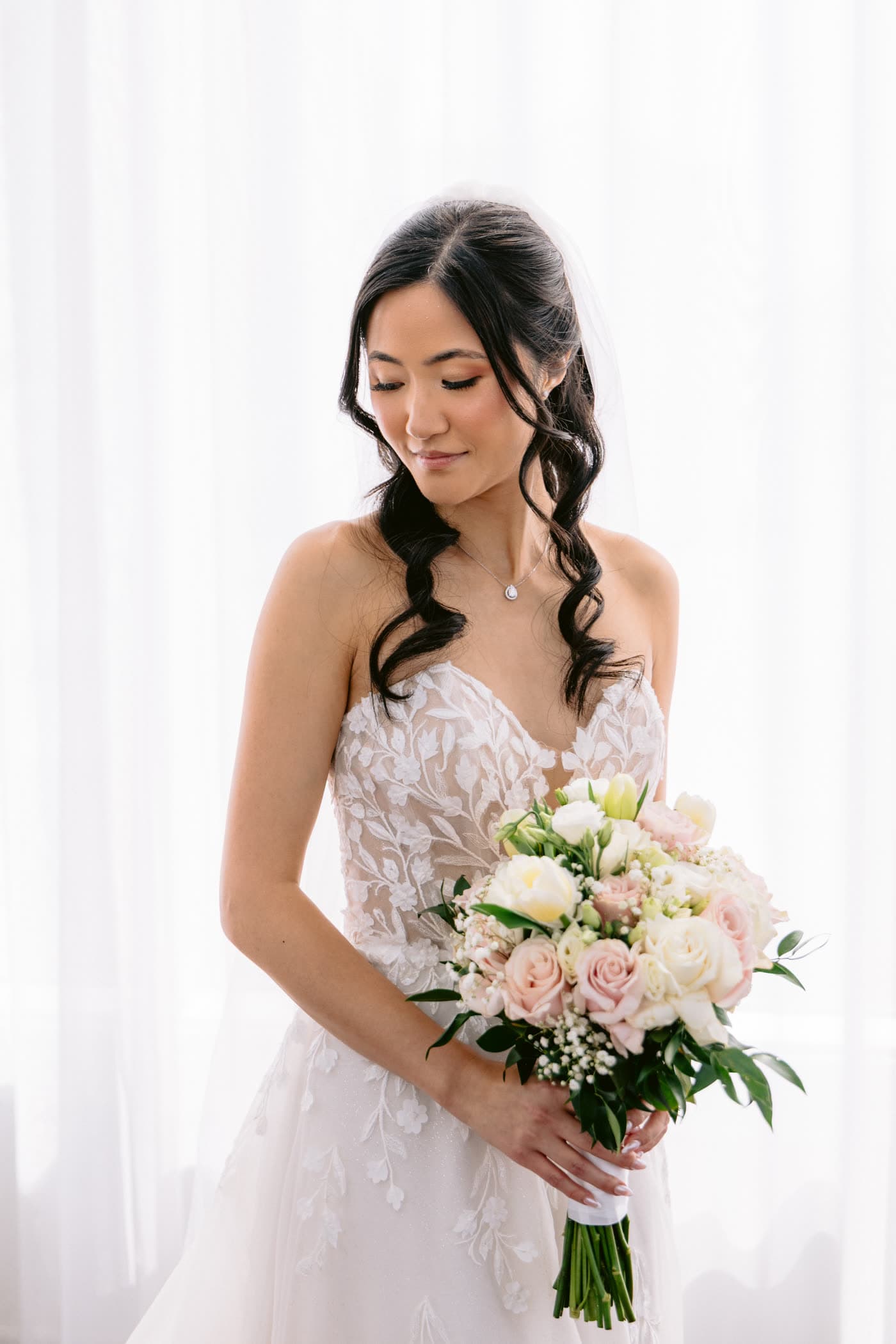 Mariée en robe de dentelle devant un voilage, bouquet de roses pâles et voile long